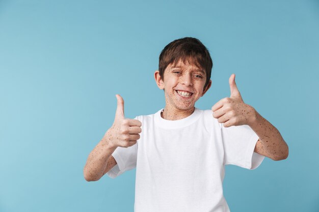 Primer retrato de feliz hermoso niño con pecas vistiendo camiseta blanca casual sonriendo y mostrando los pulgares para arriba aislado sobre la pared azul