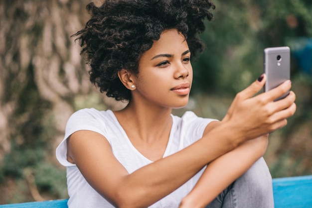 Primer retrato de una hermosa mujer afroamericana sentada al aire libre en el parque tomando selfie y enviando mensajes a través de las redes sociales usando un teléfono inteligente usando el concepto inalámbrico gratuito People and lifesetyle