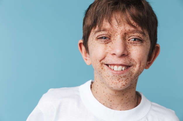 Primer retrato de un hermoso muchacho joven con pecas vistiendo camiseta blanca casual sonriendo al frente aislado sobre la pared azul