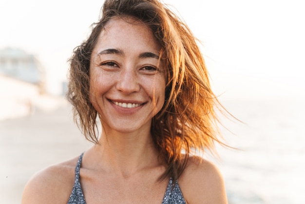 Primer retrato de mujer alegre vestida con ropa de verano sonriendo mientras camina por la playa en la mañana