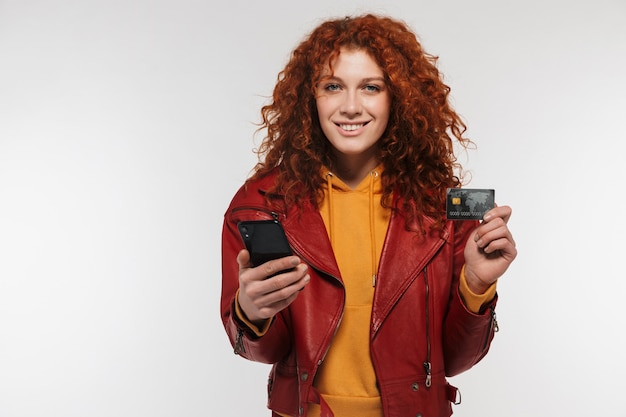 Primer retrato de mujer pelirroja feliz 20s vistiendo chaqueta de cuero sosteniendo teléfono móvil y tarjeta de crédito de plástico aislado sobre pared blanca