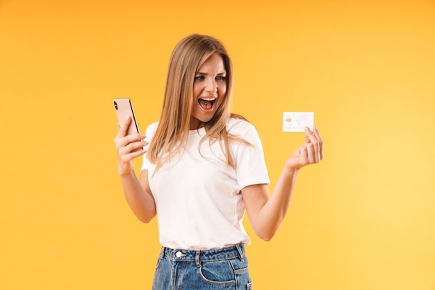 Primer retrato de mujer rubia alegre vistiendo camiseta casual gritando mientras sostiene el teléfono inteligente y la tarjeta de crédito aislado sobre la pared amarilla