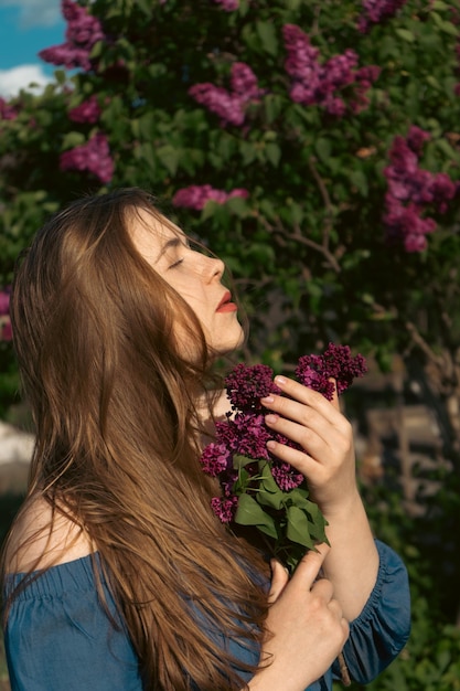 Primer retrato romántico de mujer joven en floreciente jardín lila Historia de primavera La mujer de cabello castaño sonríe con una flor en el pelo cerrando los ojos y disfrutando del estado de ánimo primaveral