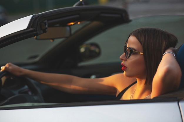 Primer tiro de modelo morena de lujo con labios rojos con gafas posando en el cabriolet