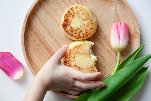 Primer tulipán rosado fresco cerca de la placa redonda de madera, la mano del niño toma dos pasteles de queso fritos mordidos de oro.