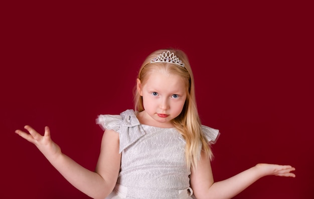 Princesa loira pequena bonita dançando no vestido branco e prata de luxo isolado em fundo vermelho. cara engraçada, emoções diferentes