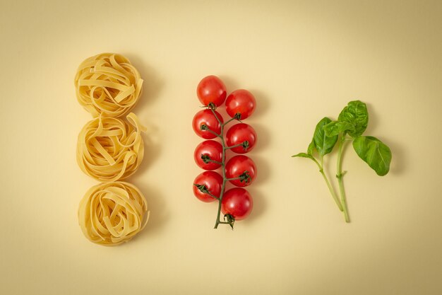 Principales ingredientes tradicionales de la cocina italiana: tagliatelle de pasta, tomates cherry y albahaca verde fresca. Concepto de comida mínima de la cocina italiana, fondo pastel amarillo claro. Vista superior, endecha plana