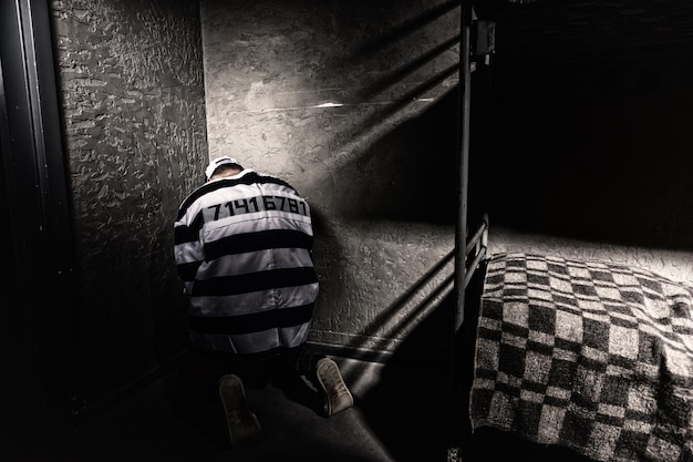 Prisioneiro vestindo uniforme de prisão com número costurado está sentado em um canto em uma pequena cela de prisão