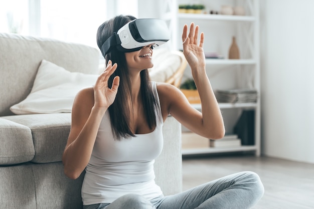 Probando su nuevo visor de realidad virtual. Mujer joven atractiva en auriculares VR gesticulando y sonriendo mientras está sentado en casa