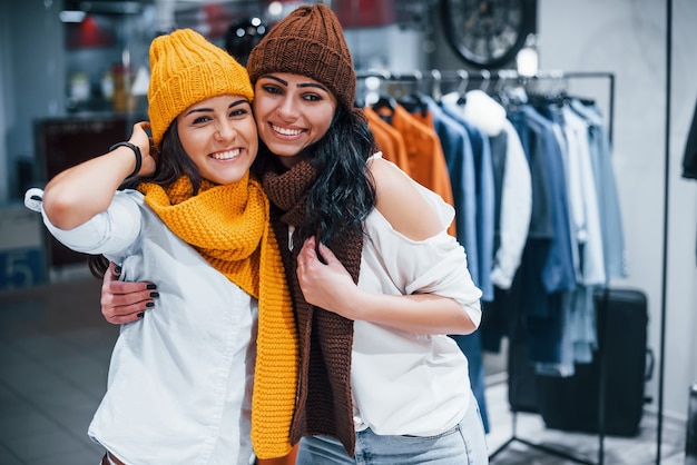 Probieren Sie warme Mützen und Schals aus. Zwei junge Frauen verbringen gemeinsam einen Einkaufstag im Supermarkt.