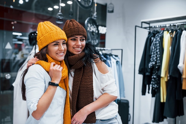 Probieren Sie warme Mützen und Schals aus. Zwei junge Frauen verbringen gemeinsam einen Einkaufstag im Supermarkt.