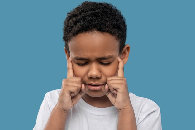 Problema, dolor. Niño de piel oscura en edad escolar primaria con los ojos cerrados y mueca infeliz tocando los dedos a la cabeza