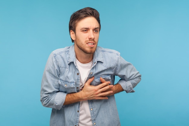 Problemas cardíacos. Retrato de homem estressado e preocupado em camisa jeans segurando o peito, sofrendo ataque cardíaco, precisa de emergência de infarto agudo do miocárdio. tiro de estúdio isolado em fundo azul