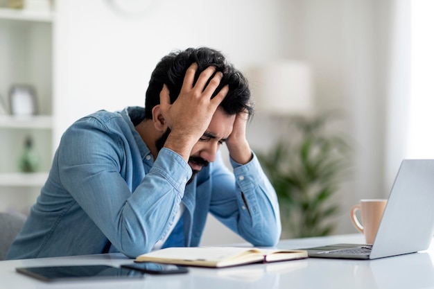 Problemas de trabalho freelancer indiano sentado na mesa tocando a cabeça em desespero