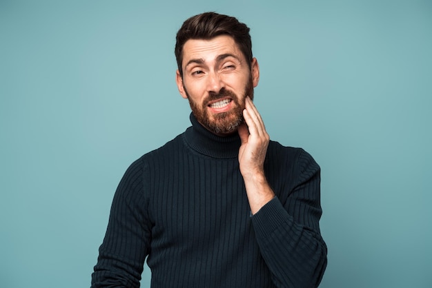 Problemas dentales Retrato de un hombre malsano que presiona la mejilla dolorida que sufre dolor de muelas agudo enfermedad periodontal caries o dolor de mandíbula Foto de estudio interior aislada en fondo azul