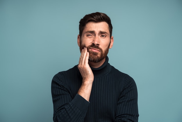 Foto problemas dentários. retrato de homem insalubre pressionando a bochecha dolorida, sofrendo de dor de dente aguda, doença periodontal, cáries ou dor na mandíbula. foto de estúdio interna isolada em fundo azul