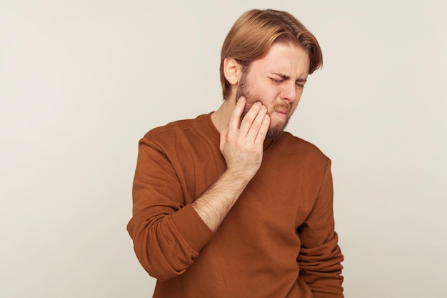 Problemas de salud dental Retrato de hombre barbudo en sudadera con dolor de mejilla frunciendo el ceño por dolor de dientes agudo