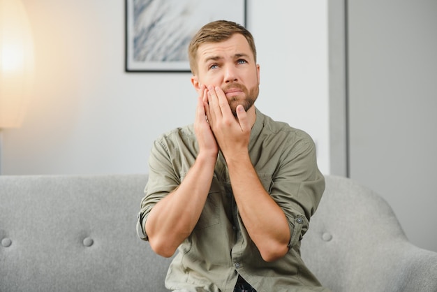 Probleme mit Zähnen Zahnschmerzen Verärgerter Kaukasier sitzt zu Hause auf der Couch und hält seine Hand an seine Wange mit Zahnschmerzen braucht eine Zahnarztberatung trauriger Gesichtsausdruck