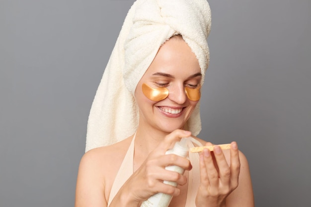 Foto procedimentos cosméticos cuidados com a pele jovem alegre feliz enrolada na toalha com manchas sob os olhos usando loção para cuidar da pele do rosto posando isolada sobre fundo cinza