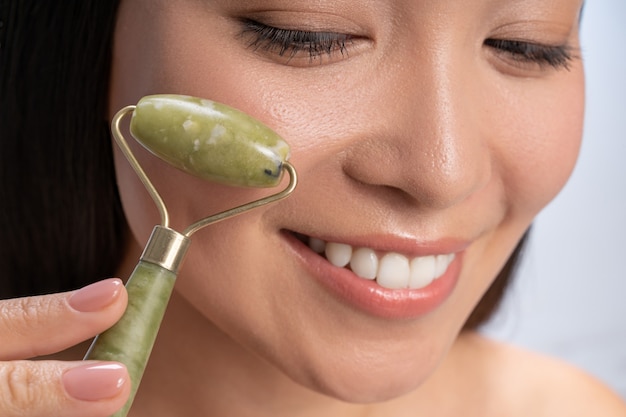 Procedimiento de belleza. Linda mujer joven con rodillo de jade y demostrando una sonrisa amistosa durante el procedimiento