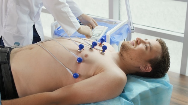 Foto procedimiento de electrocardiograma para diagnosticar enfermedades cardíacas. un cardiólogo coloca electrodos en el pecho desnudo de un joven acostado en el sofá para tomar un electrocardiograma en el consultorio de la clínica.