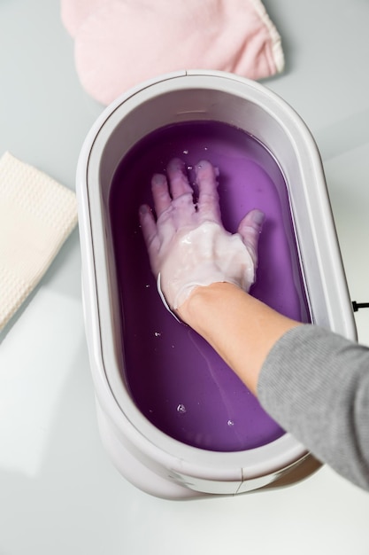 Procedimiento de toma de manos femeninas en un recipiente de cera de parafina lila. Equipo cosmetológico y de cuidado de la piel en un salón de belleza y spa.