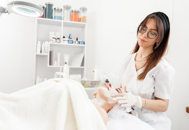 Foto procedimientos cosmetológicos el médico del salón de belleza con guantes aplica una máscara para una mujer hermosa con los ojos cerrados