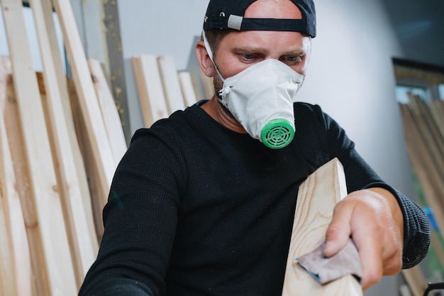 Procesamiento de madera para la fabricación de muebles Un carpintero con ropa oscura y un respirador muele tablas con papel de lija