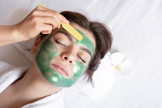 El proceso de aplicar una mascarilla cosmética verde en el rostro de una mujer joven.