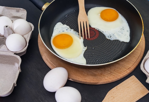 Proceso de cocción de huevos fritos. Dos huevos fritos en una sartén y huevos crudos sobre una madera oscura.