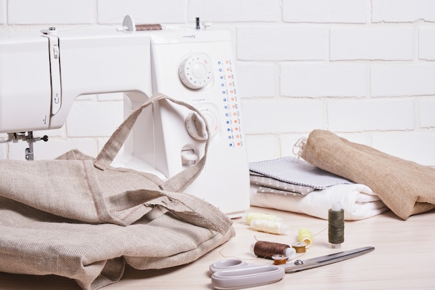 El proceso de coser bolsas ecológicas, hilo, tijeras y una máquina de coser en el fondo de un espacio de copia de pared de ladrillo blanco