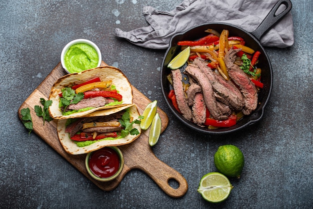 Proceso de elaboración de un plato tradicional mexicano Tacos de fajitas de carne servidos en una tabla de cortar de madera con salsa de tomate y guacamole sobre fondo de piedra rústica desde arriba, comida mexicana estadounidense