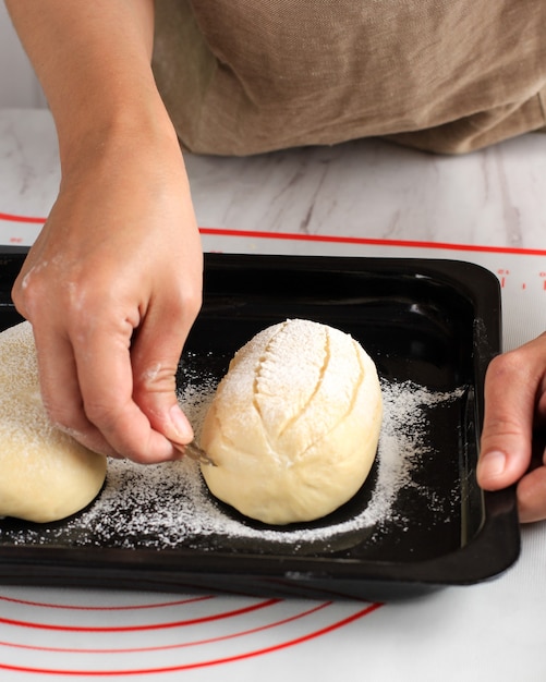 Foto proceso de horneado: hacer pan, puntuar la masa de pan de hogar de leche japonesa redonda cruda. mano femenina haciendo un corte transversal en la parte superior de la masa con una maquinilla de afeitar