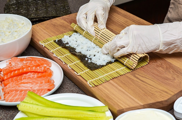 Proceso de preparación de sushi. Las manos de las mujeres en guantes de goma envuelven rollos para sushi, sushi en casa