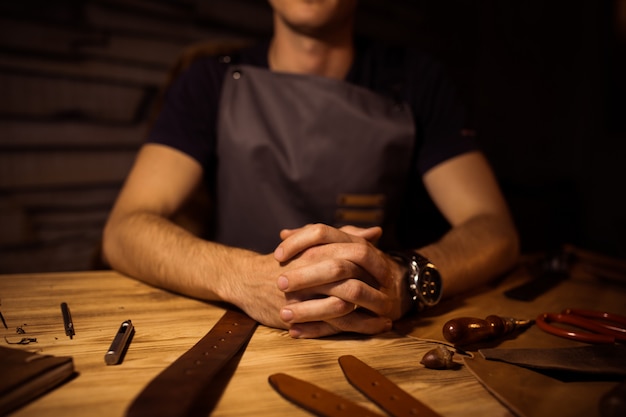 Proceso de trabajo del cinturón de cuero en el taller de cuero. Hombre de la mano en la mesa de madera. Elaboración de herramientas en el fondo. Curtidor en la antigua curtiduría. Ciérrese encima del brazo de los hombres.