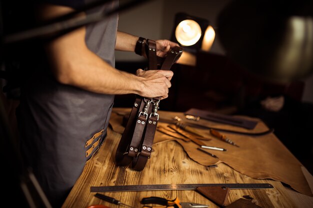 Proceso de trabajo del cinturón de cuero en el taller de cuero. Hombre sujetando el cinturón del fotógrafo para la cámara. Herramienta sobre fondo de madera. Curtidor en la antigua curtiduría.