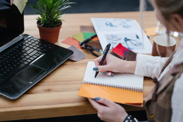 Proceso de trabajo del diseñador de moda. escritorio de oficina con bocetos de muestras de color y suministros sobre la mesa. mujer escribiendo notas e ideas en su bloc de notas