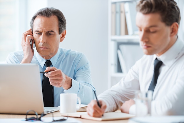 Proceso de trabajo. Hombre de negocios serio hablando por teléfono móvil y apuntando a la computadora portátil