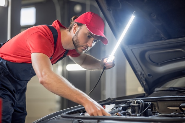 Proceso de trabajo. Joven mecánico con barba en camiseta roja y gorra con lámpara que controla la anatomía del coche debajo del capó