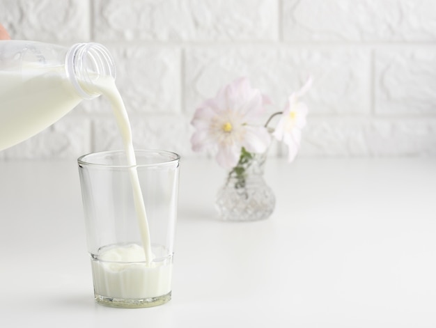 Proceso de vertido de leche fresca de una botella de plástico en un vaso de vidrio, mesa blanca