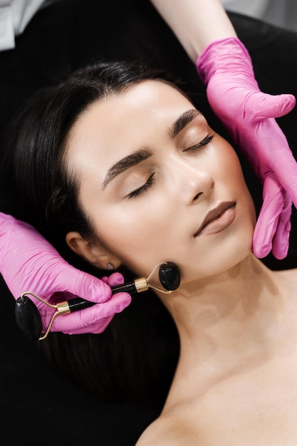 Processo de massagem facial com rolo de pedra preta closeup Cosmetologista usando rolo de pedra para massagem facial para paciente menina Massagem facial com rolo de cosméticos na clínica de cosmetologia