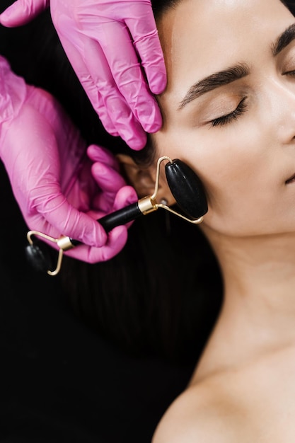 Foto processo de massagem facial com rolo de pedra preta closeup cosmetologista usando rolo de pedra para massagem facial para paciente menina massagem facial com rolo de cosméticos na clínica de cosmetologia