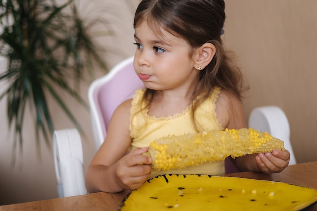 Processo de menina comendo milho fresco em casa adorável criança se senta na mesa e come milho