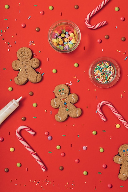 Processo de preparação do biscoito de gengibre de Natal com homem-biscoito, glacê, granulado de açúcar e bastões de doces em um fundo de mesa vermelho. Plano de papel de parede de ano novo lay.
