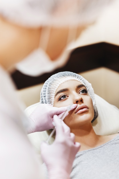 Foto processo de preparação para o aprimoramento labial. o médico retira ácido hialurônico na frente do paciente. a jovem com um rosto bonito no chapéu especial e mãos do médico na luva rosa