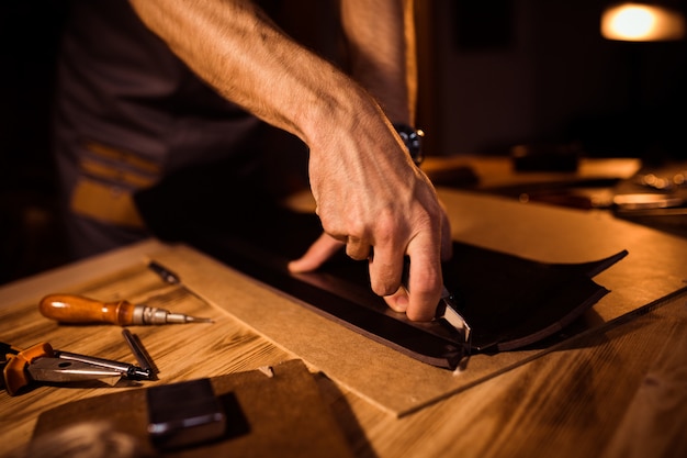 Processo de trabalho do cinto de couro na oficina de couro. homem segurando a ferramenta de elaboração e trabalho. curtidor em curtume antigo. fundo de mesa de madeira.