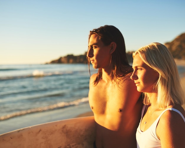 Procurando a onda perfeita Foto recortada de um jovem casal com uma prancha de surf juntos e assistindo o pôr do sol na praia