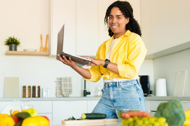 Procurando novas receitas Mulher negra feliz segurando e usando laptop enquanto cozinha salada fresca na cozinha moderna