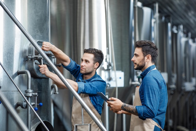 Produção de cerveja artesanal na fábrica. Fermentação de bebida, pequena empresa familiar e produção cervejeira. Jovens bonitos sérios em aventais torcem ventel em tanque de metal para cerveja lager, espaço vazio