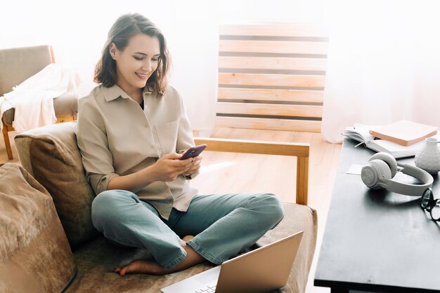 Foto productiva millennial workday mujer sonriente mira el teléfono lee mensajes multitarea con la computadora portátil